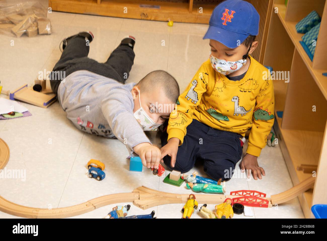 Education Preschool 3-4 year olds two boys playing together in block area, playing with train set and people figures, wearing face masks Covid-19 Stock Photo