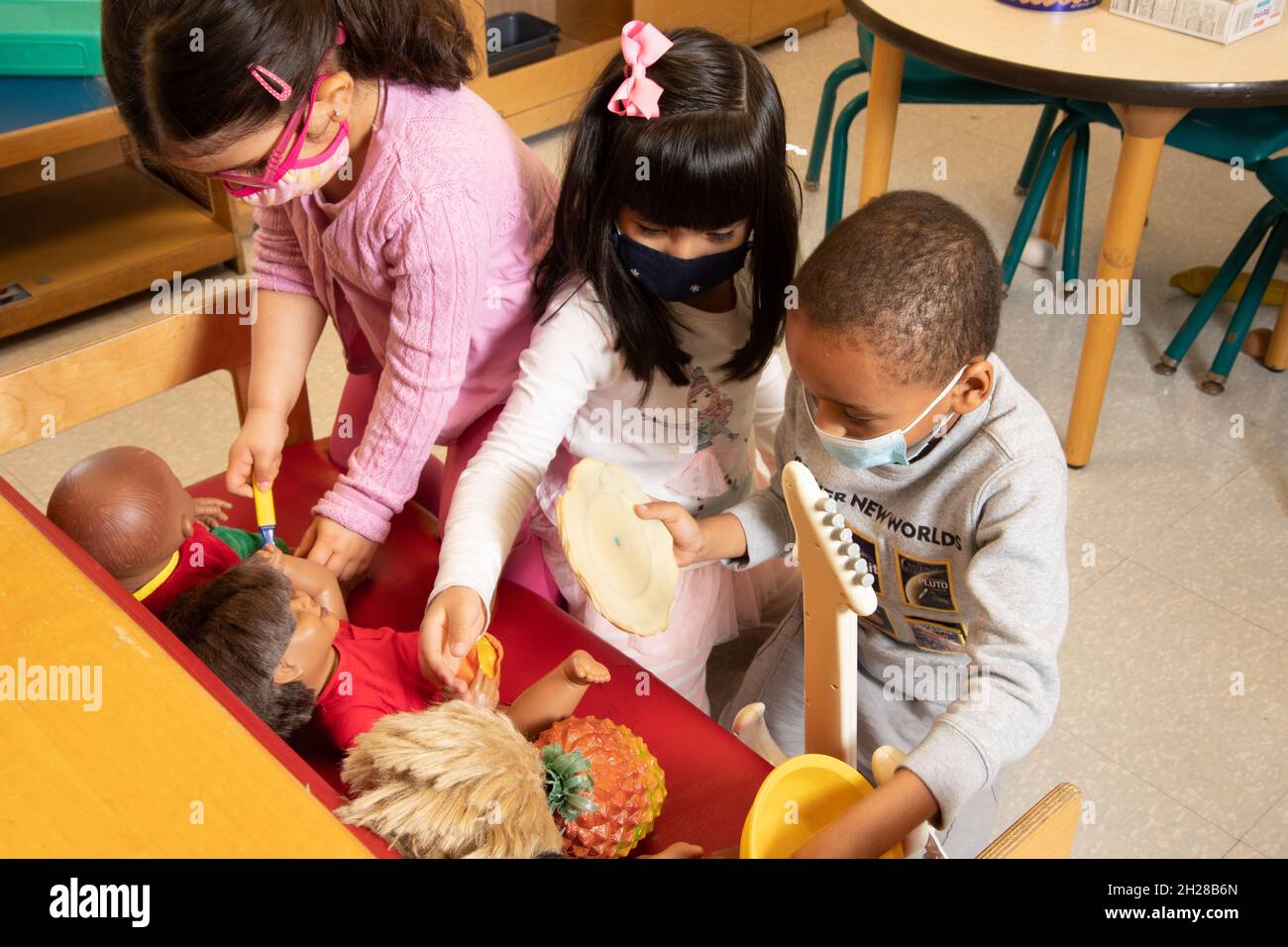 Education Preschool 3-4 year olds two girls and a boy playing with dolls and toy food in family area, wearing face masks to protect against Covid-19 Stock Photo