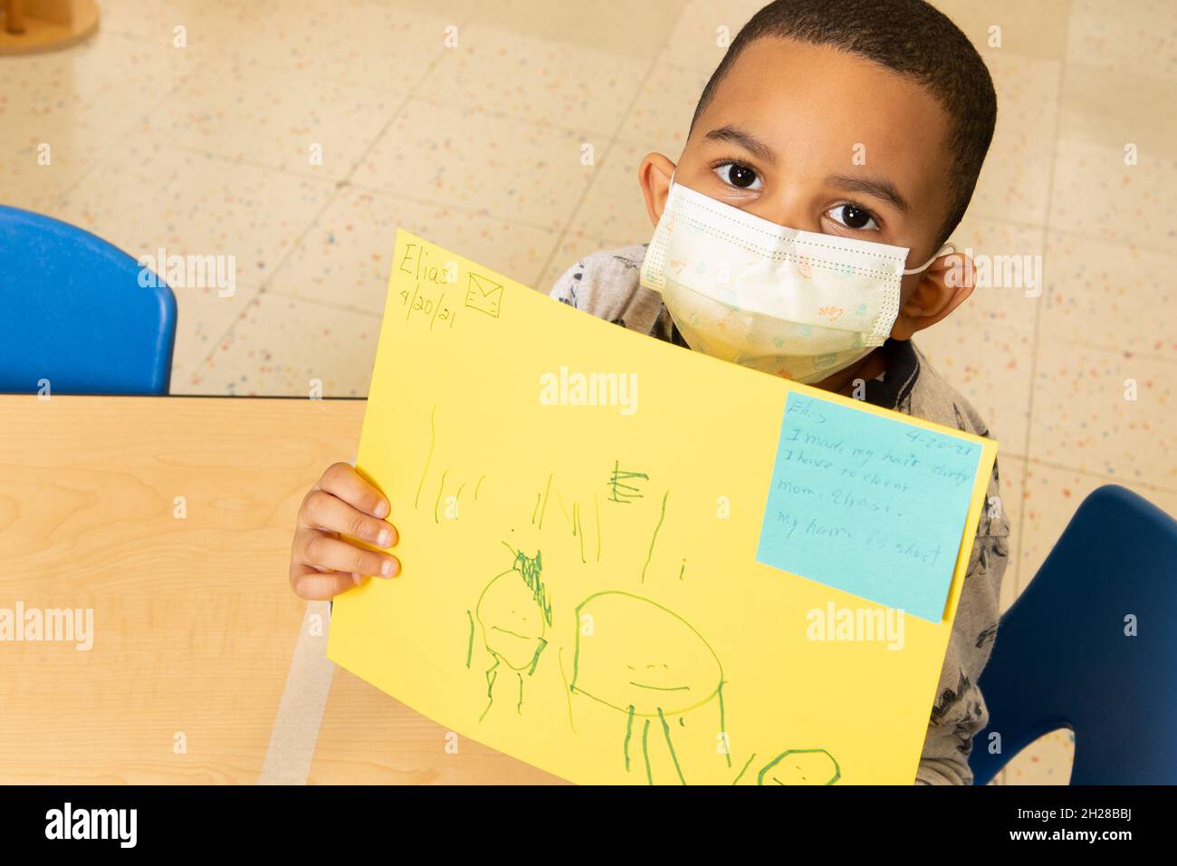 Education Preschool 4-5 year olds boy holding up drawing he did of his family, wearing face mask to protect agains Covid-19 infection Stock Photo