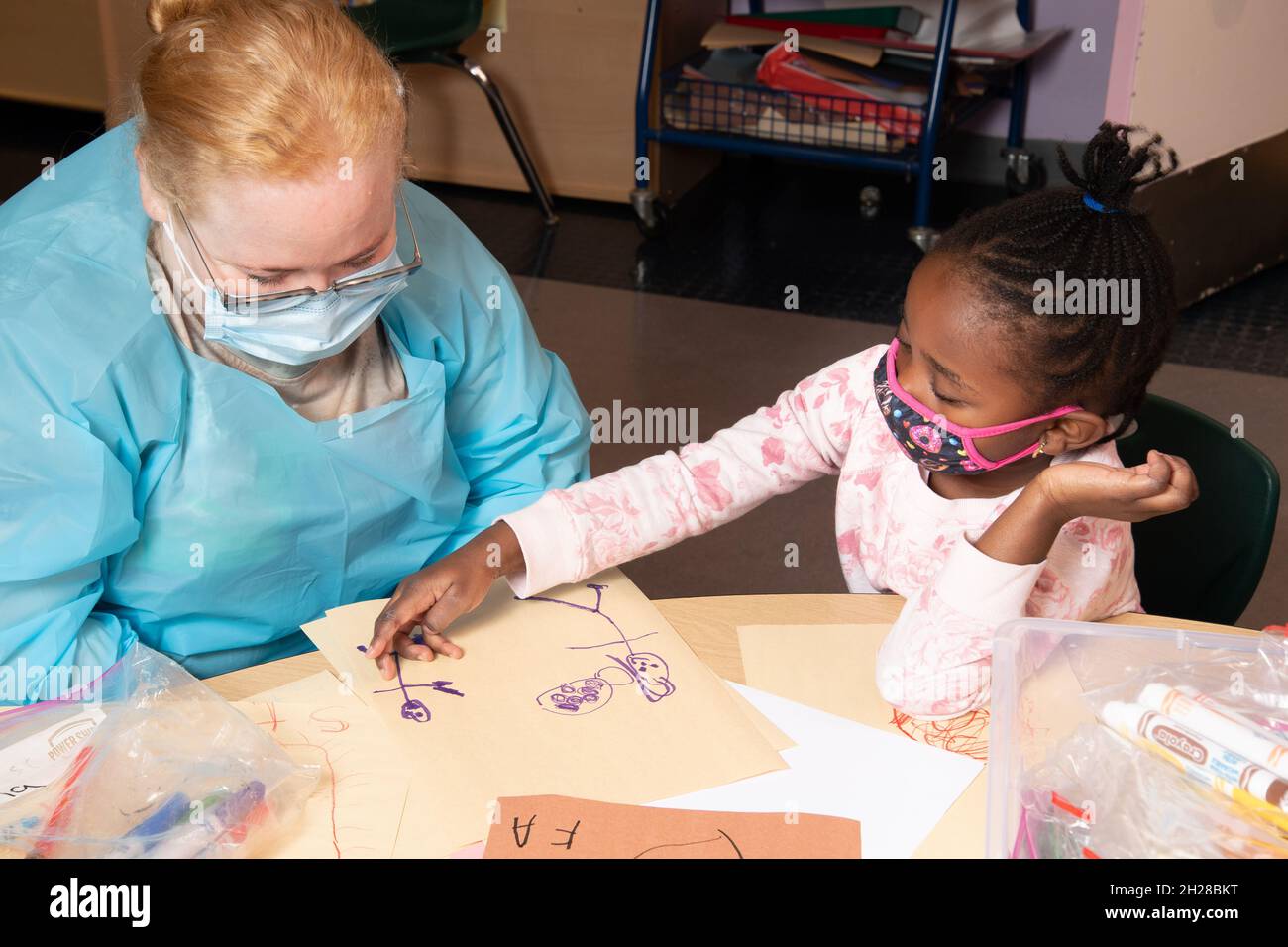 Education Preschool 4-5 year olds girl telling teacher about her drawing describing what is in the picture, both wearing face masks Covid-19 Stock Photo