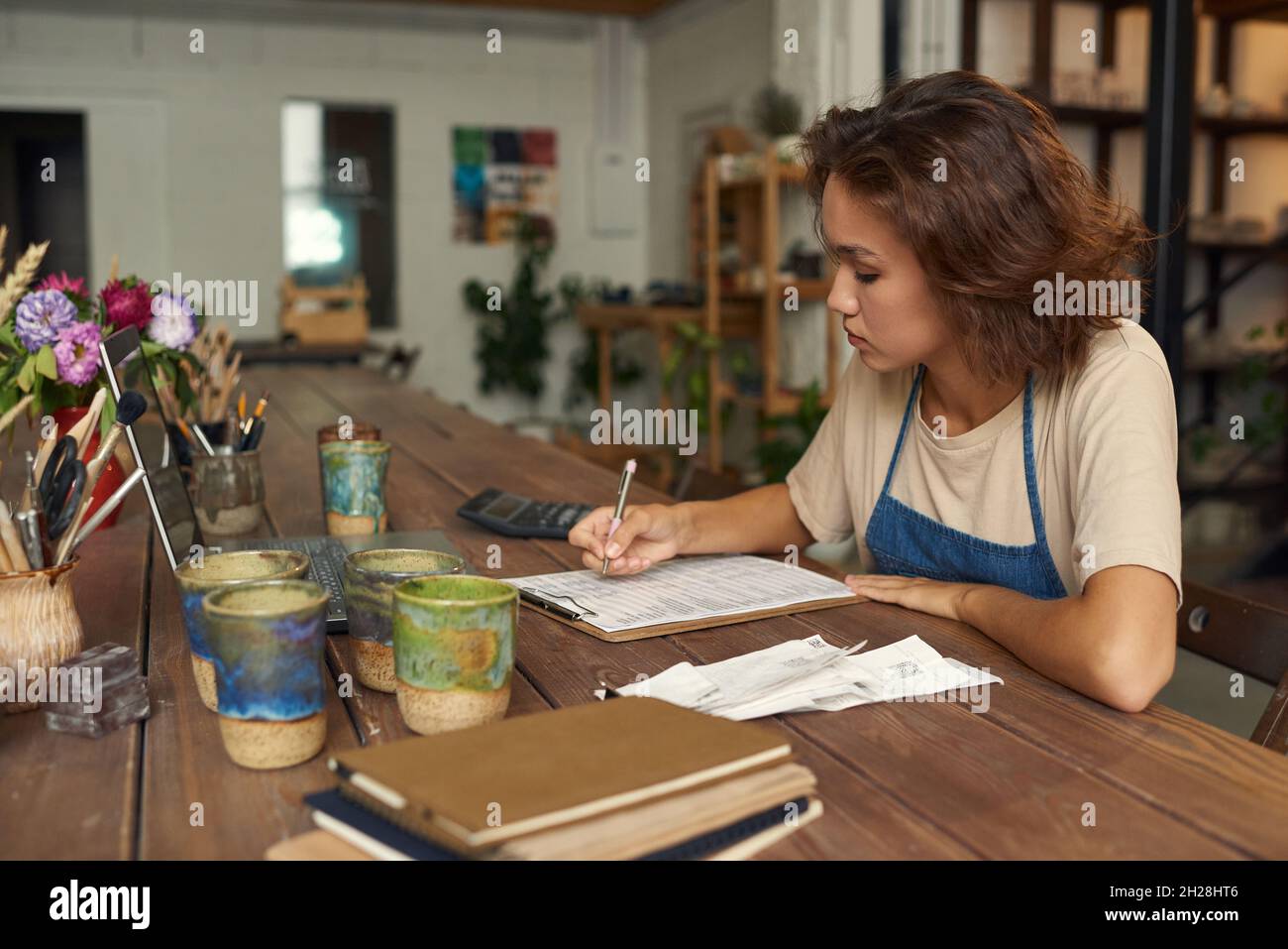 Concentrated young woman sitting at wooden table with ceramic mugs and ananlyzing receipts while working in pottery workshop Stock Photo