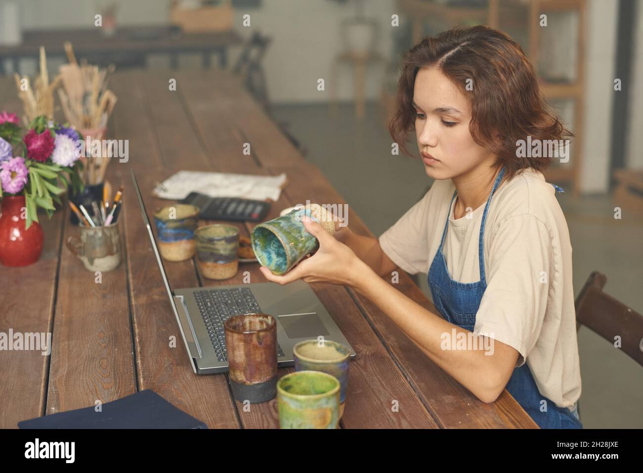 Serious young Caucasian woman sitting at wooden table with laptop and examining ceramic mug in pottery workshop Stock Photo
