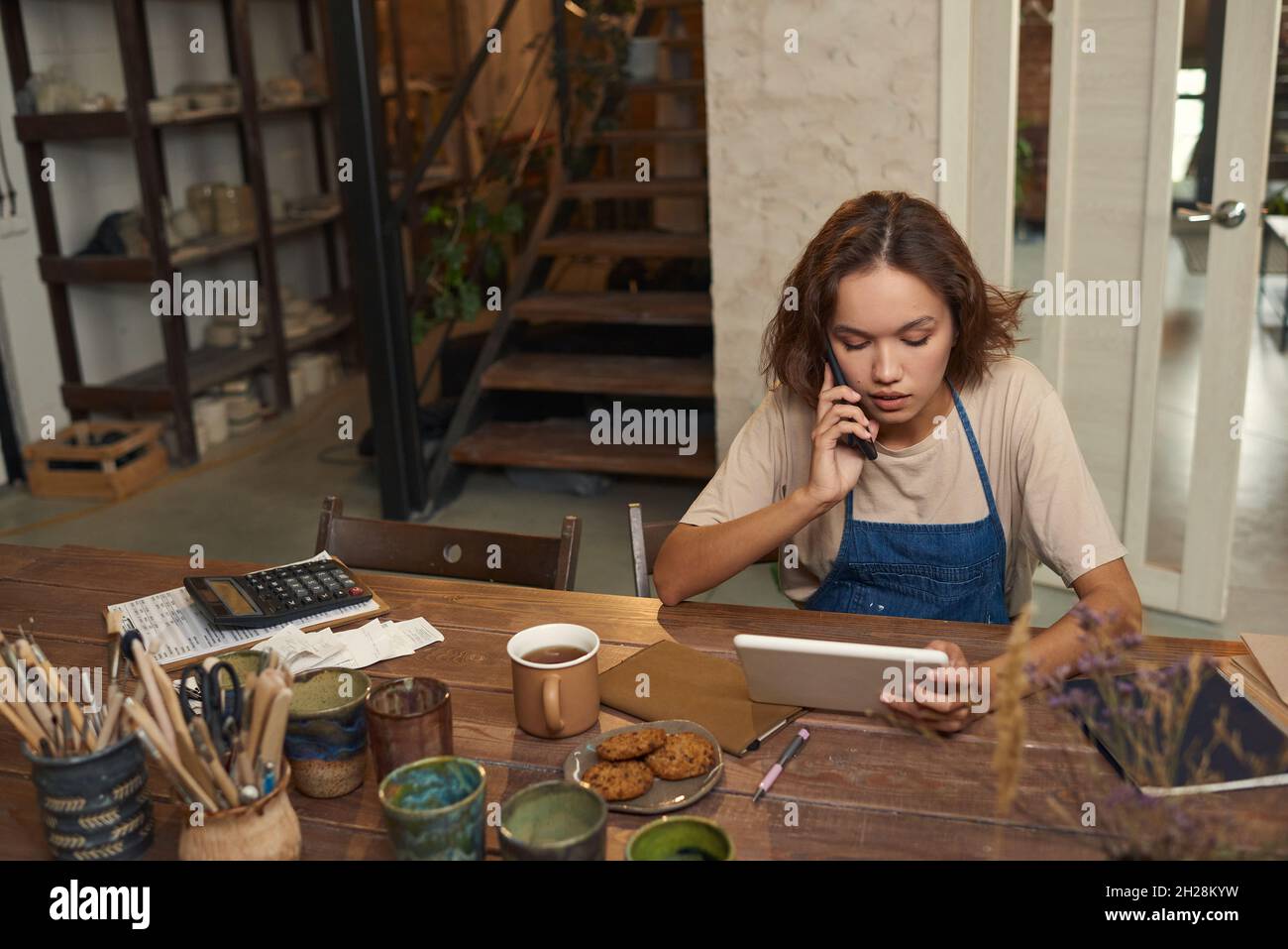 Busy young female potter sitting at desk with tools and ceramic mugs and using tablet while calling supplier in workshop Stock Photo