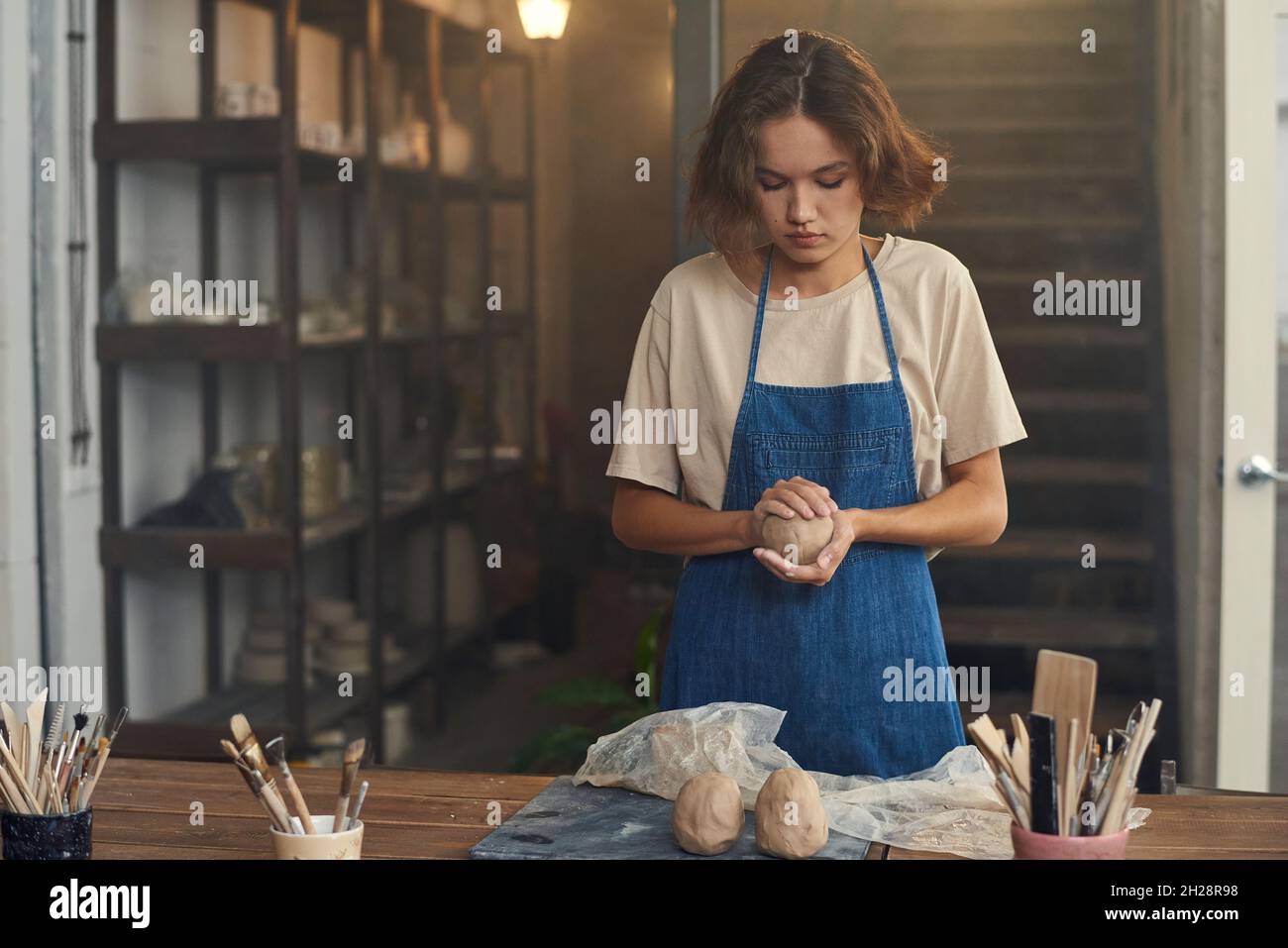 Concentrated young female potter in apron balling up clay while preparing it for sculpting in workshop Stock Photo