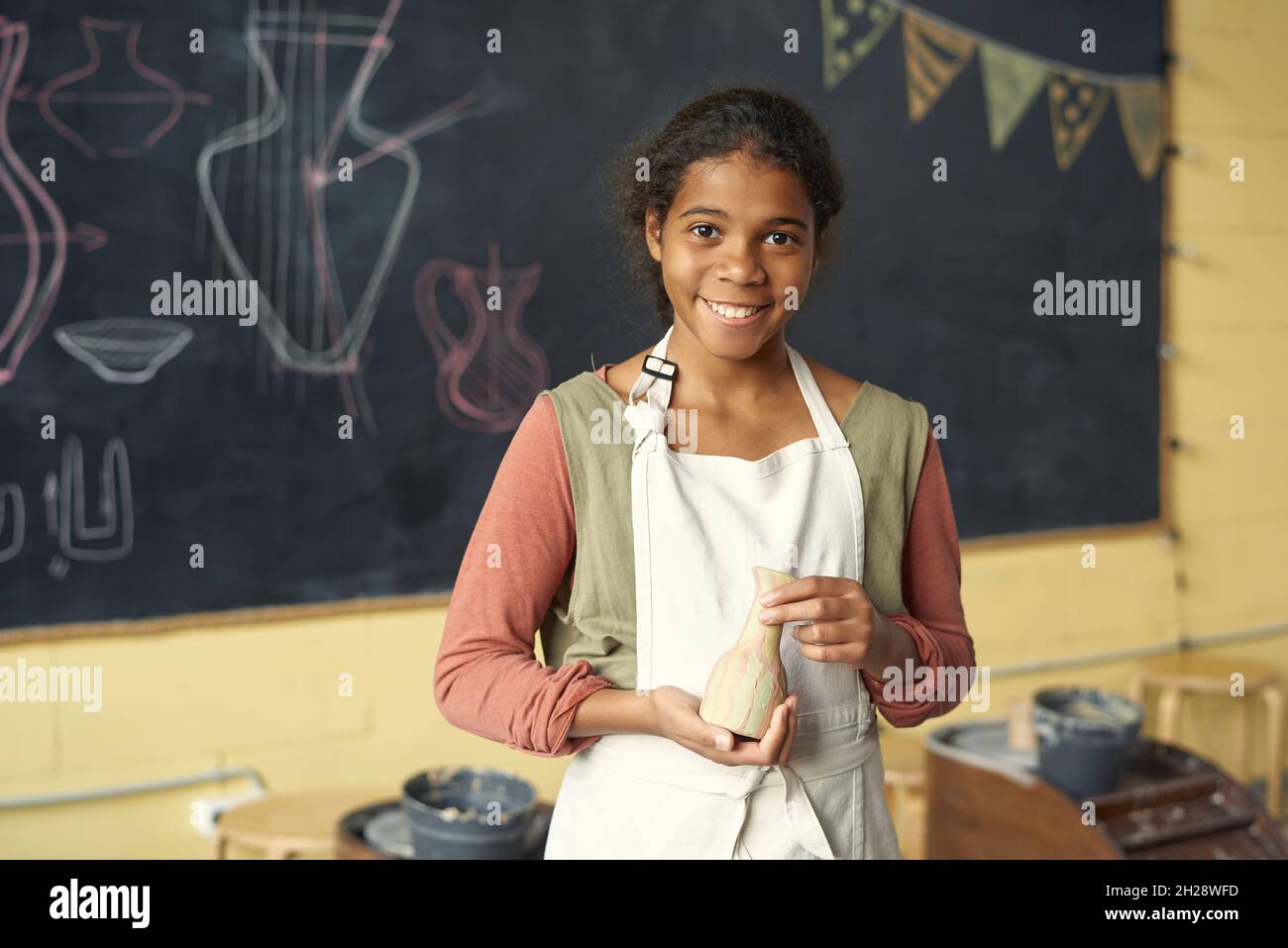 Portrait of smiling African American girl in white apron standing with handmade vase against blackboard at pottery class Stock Photo