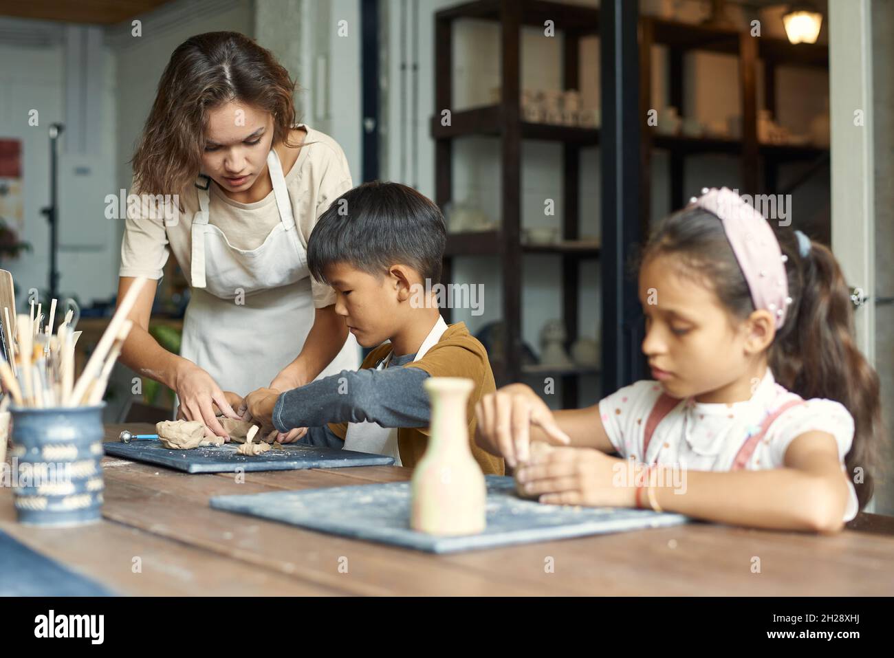 Young teacher in apron standing at Asian boy and helping him to sculpt clay at ceramics class Stock Photo