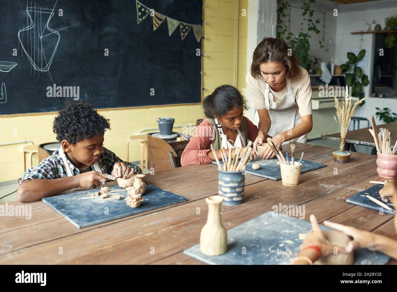 Young teacher in apron sculting with kids and assisting them at ceramics class Stock Photo