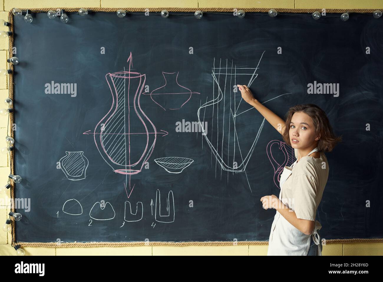 Portrait of young ceramics teacher in apron drawing vases on blackboard at pottery class Stock Photo