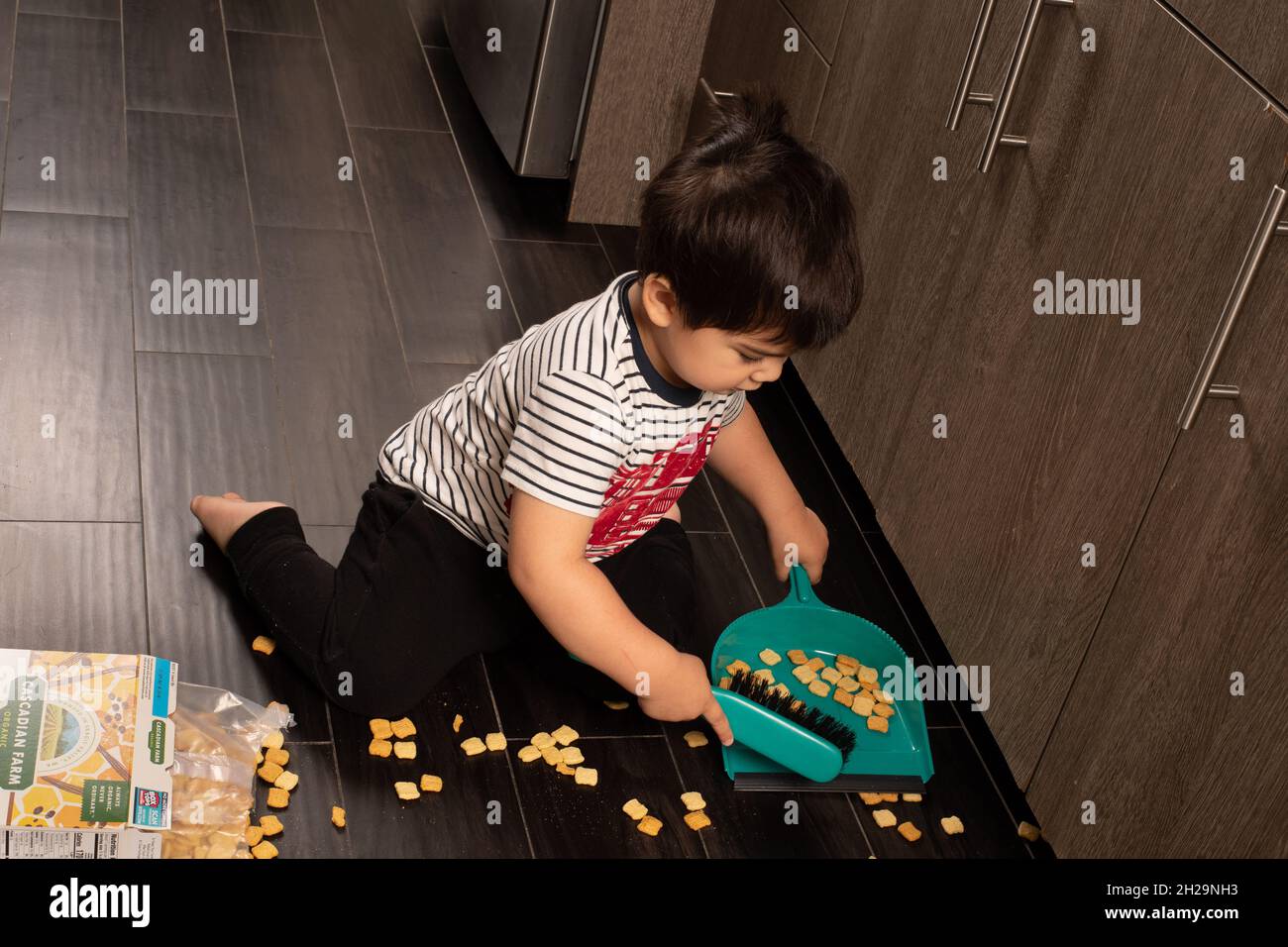 Two year old toddler boy sweeping up spilled cereal using dust pan and brush Stock Photo