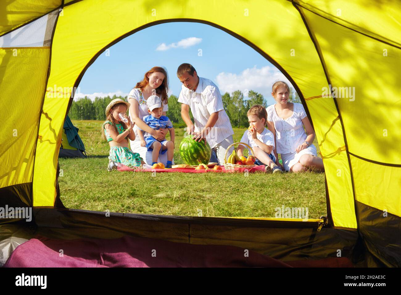 Outdoor group portrait of happy family having picnic on green grass in camp. Father is cutting watermelon.. View from camping tent entrance. Stock Photo