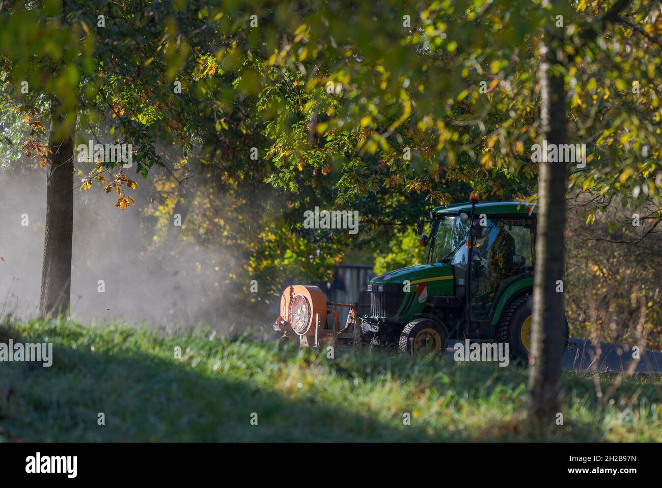 Small tractor of the municipality with front mounted leaf blower device driving through public park cleaning the walkways on sunny autumn day in Octob Stock Photo