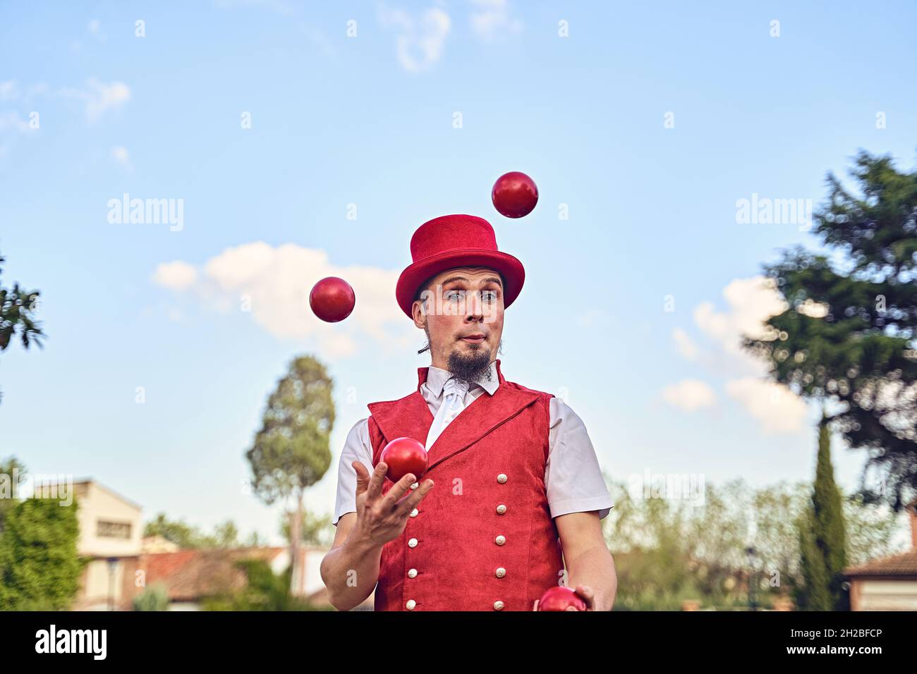 Bearded male performer in red hat and waistcoat looking at camera and juggling balls during show in park on sunny summer day Stock Photo