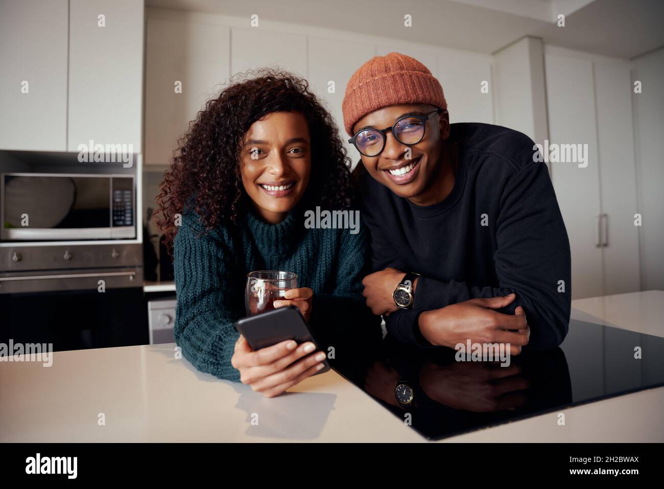 Multi-ethnic couple smiling at camera while in kitchen in modern apartment using cellphone Stock Photo