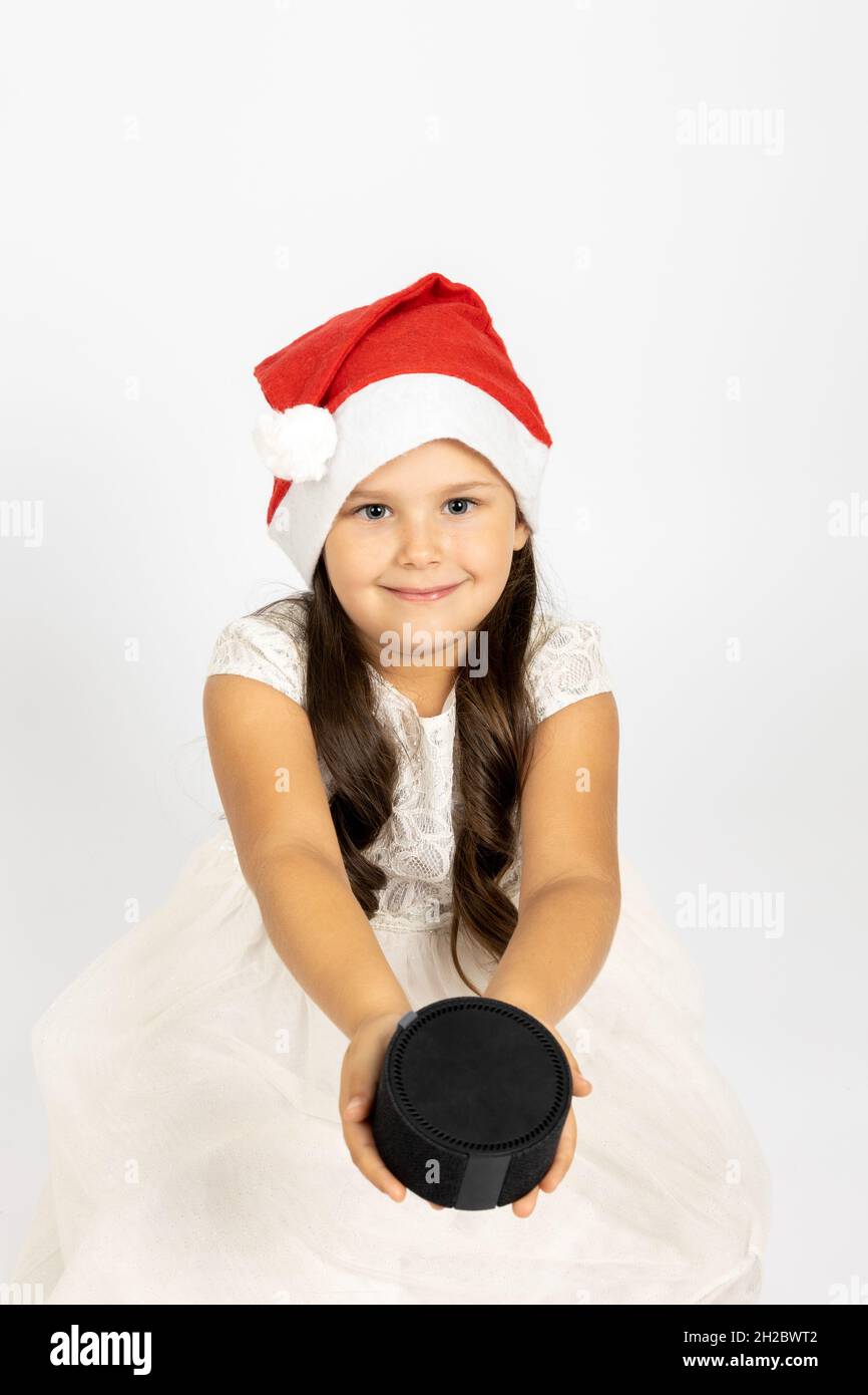portrait of smiling pre-school age girl in Santa Claus hat holding out smart music speaker or station to camera, isolated on white background, concept Stock Photo