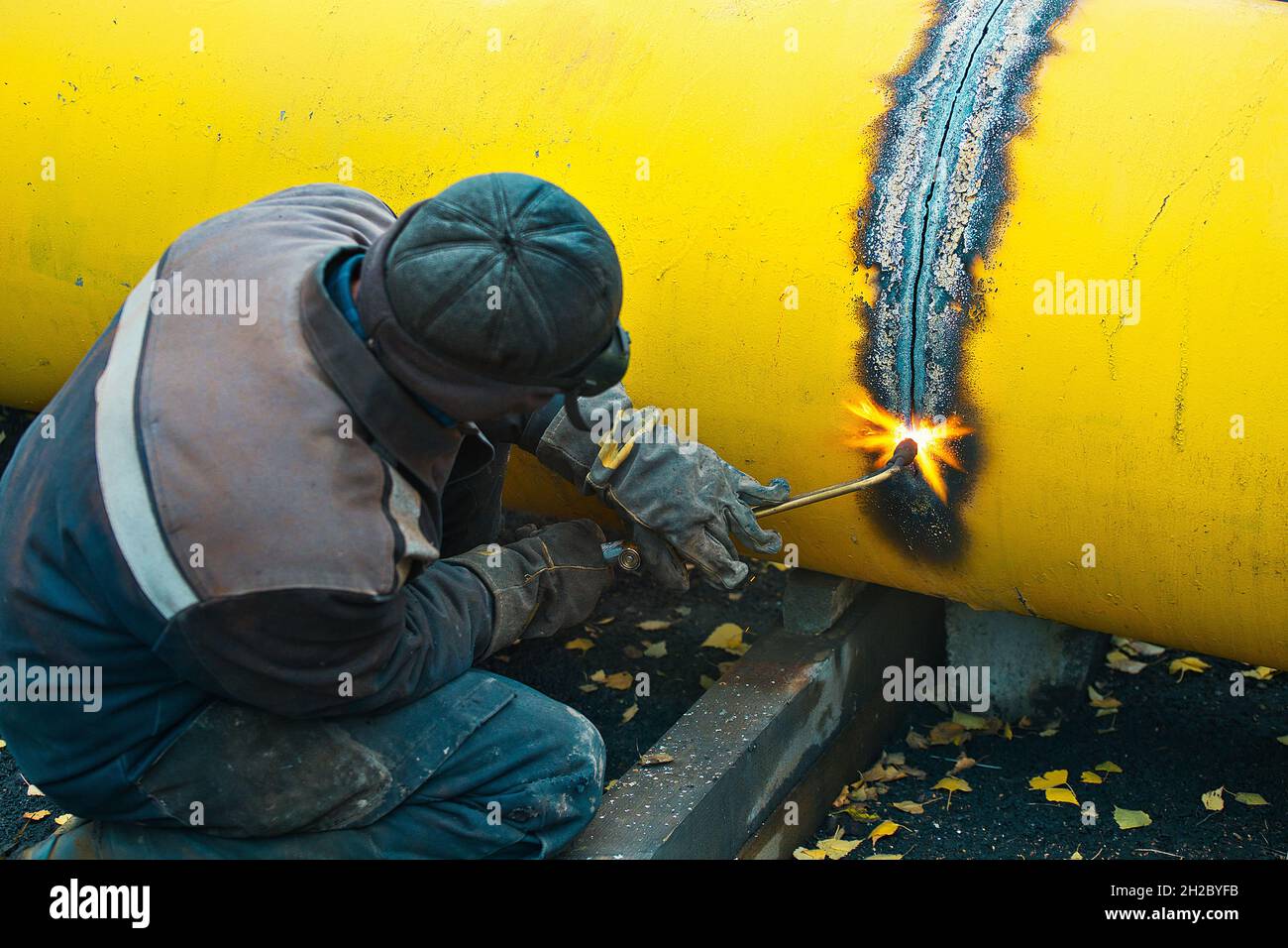 The welder cuts a large yellow pipe with acetylene welding for gasification. Disposal of old used metal pipes. Authentic workflow scene. Industrial background. Stock Photo