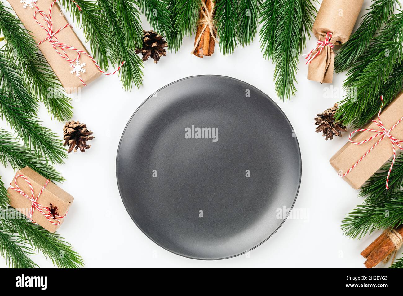 mock-up of black empty ceramic plate on white table with Christmas decorations made of fir branches, cones and gift boxes Stock Photo