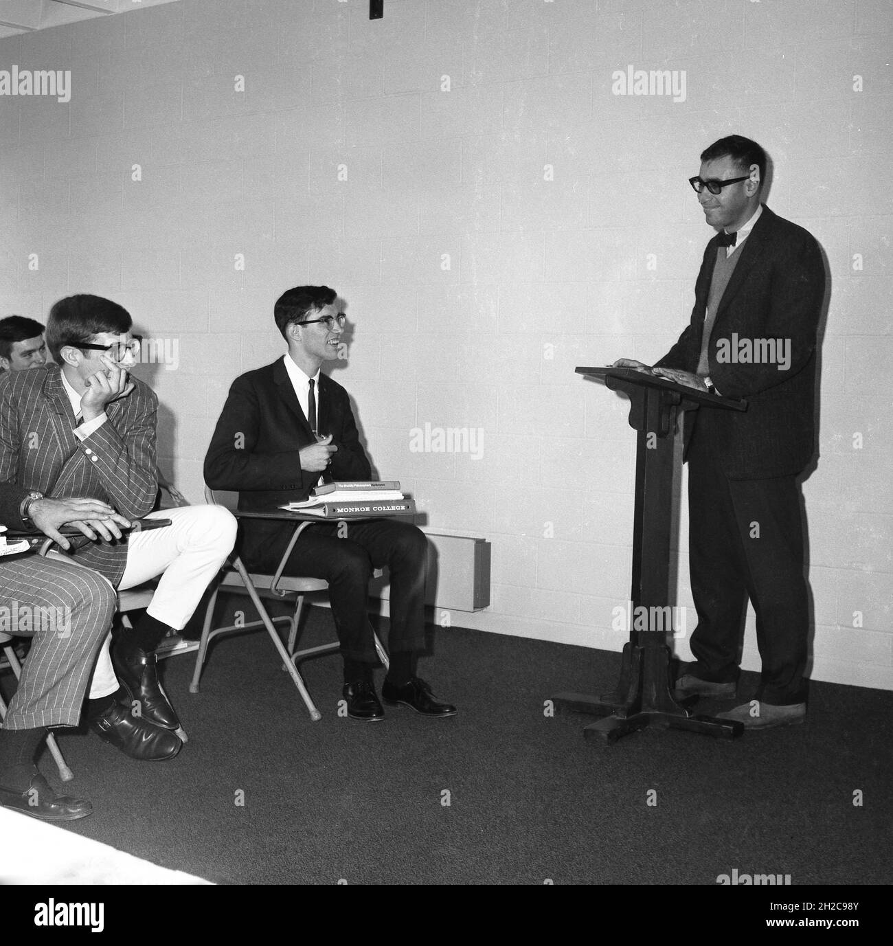 1960s, historical, young adult male students in a philosophy class sitting infront of a male teacher or lecturer standing at a lectern, USA. One of the young men has on his lap a book by Robert Heilbroner, The Worldly Philosophers. Stock Photo