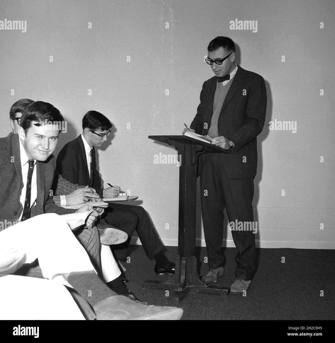 1960s, historical, an adult male teacher wearing a bowtie, suit and swede shoes standing at a lectern, reading to some male undergraduate students in a philosophy class, USA. One of the student's winks at the camera. Stock Photo