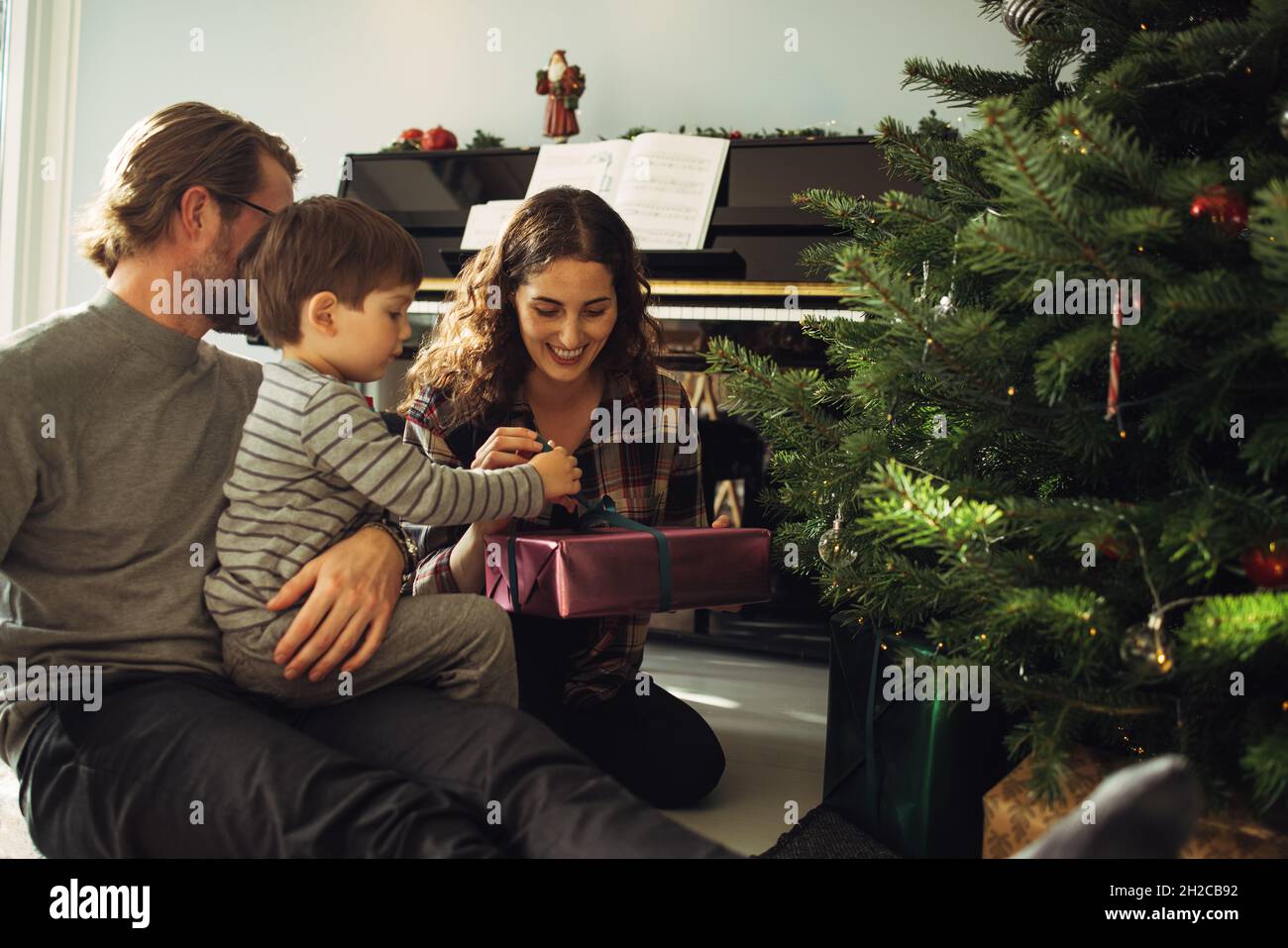 Boy opening his Christmas gift sitting with parents. Small family sitting near Christmas tree unboxing gifts. Stock Photo