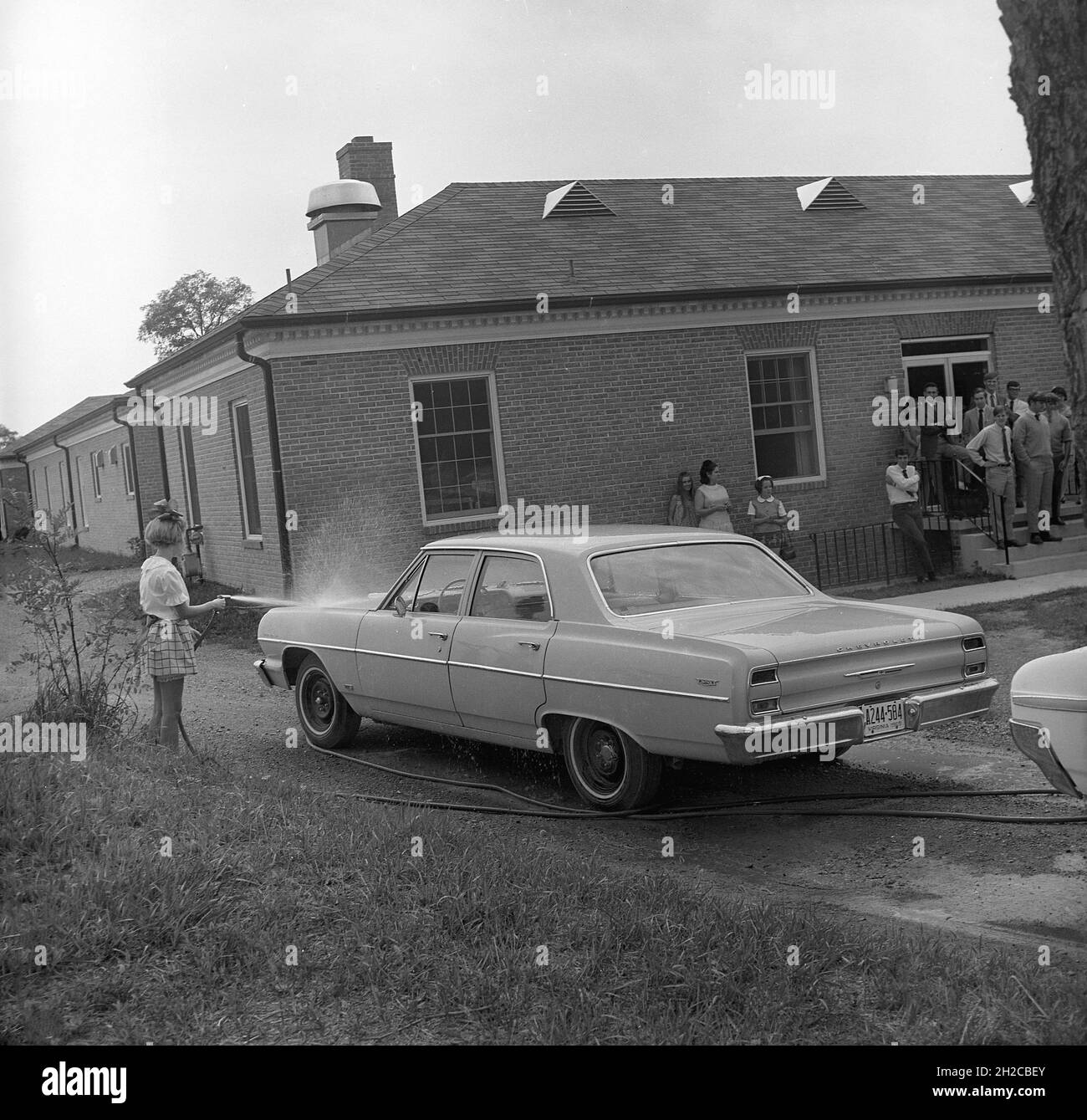 1960s, historical, outside a single-storey building at a college, with students and teachers looking on, a teenage girl in a skirt and ankle socks using a jet spray to wash an American Chevrolet automobile, Middletown, Virginia, USA. Stock Photo
