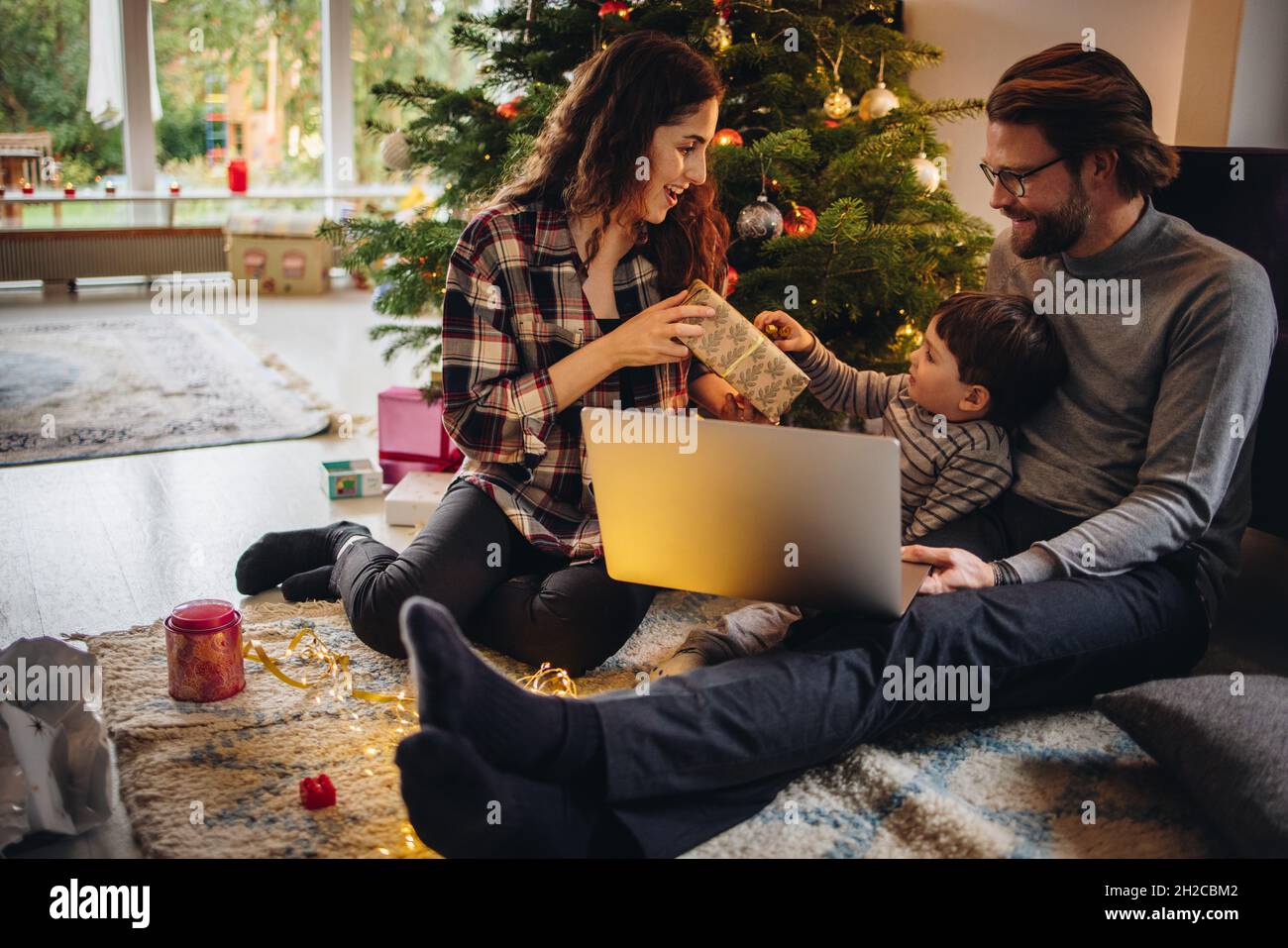 Family sitting by Christmas tree at home. Boy opening his Christmas gift while sitting with his parents. Stock Photo