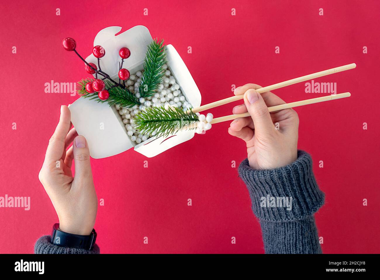 top view Christmas decorations in box for Chinese noodles. Female hands eat with chopsticks. white pearl beads, branches of Christmas tree in box on red table. Asian Chinese or Korean New Year concept Stock Photo