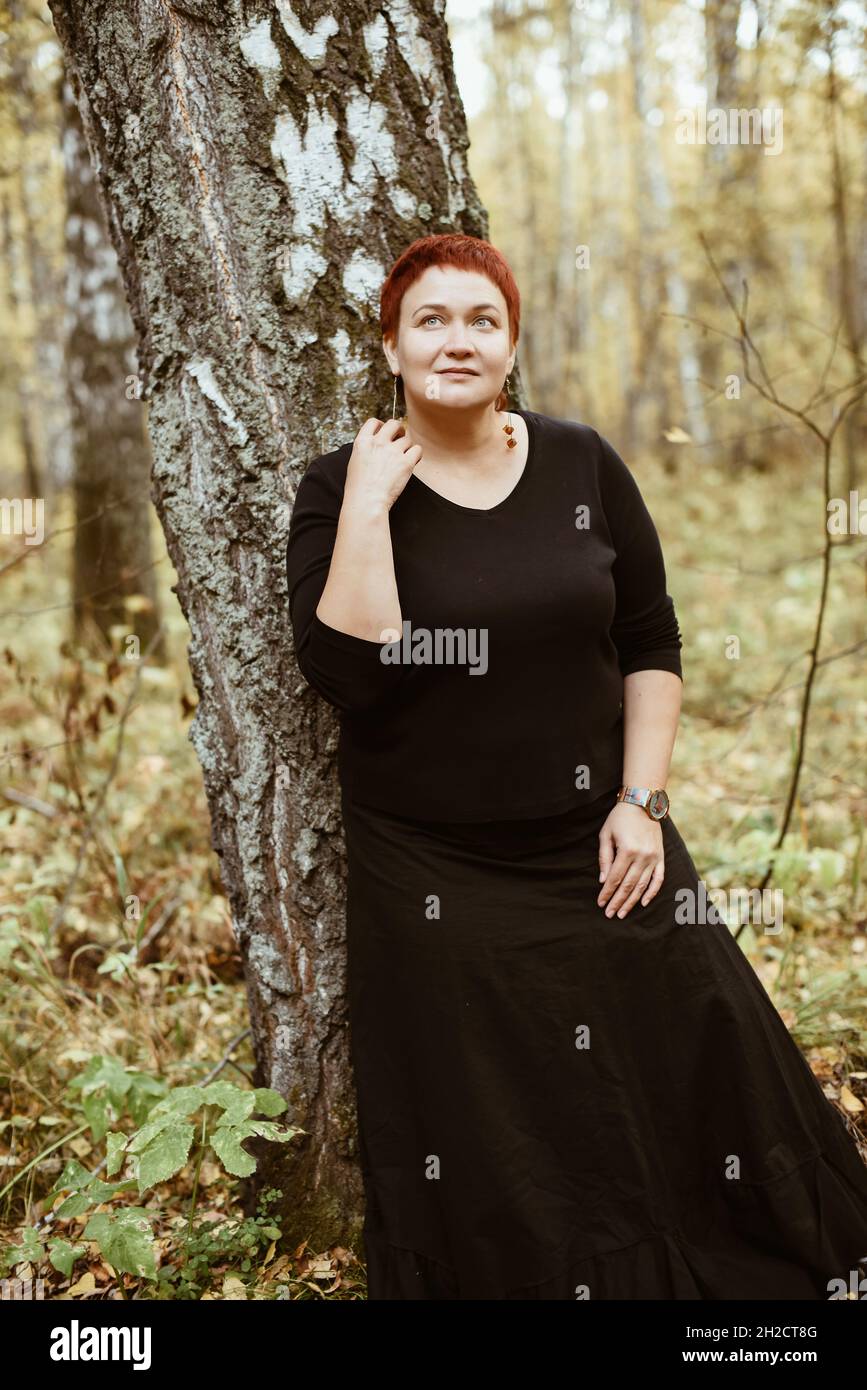 Middle-aged woman of 30-40 years old in autumn forest stands by tree, pensive, dreamy. Stock Photo