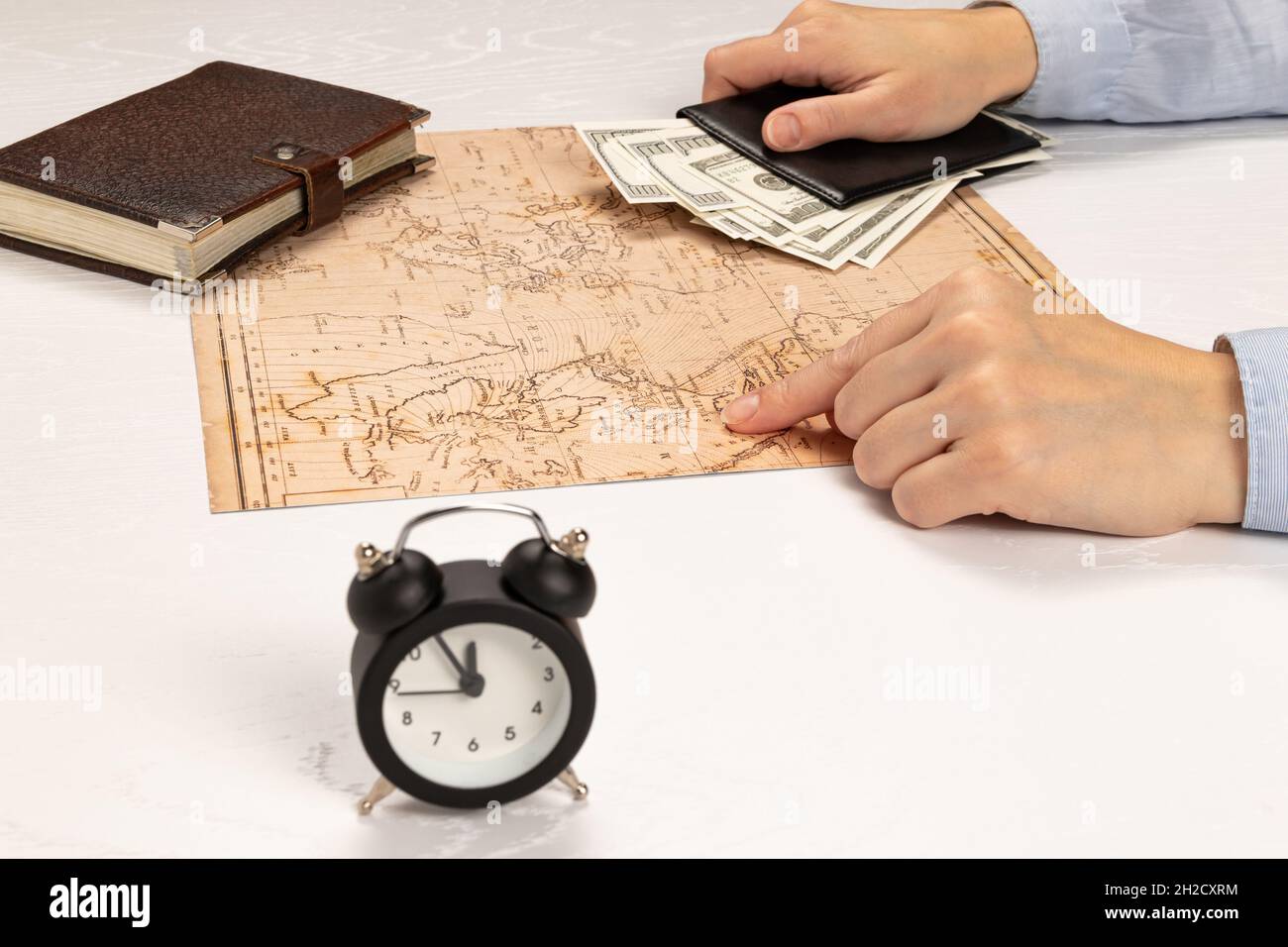 female hands hold a black leather wallet with money and show the resting place on the world map. on a white wooden table brown diary and alarm clock. Stock Photo