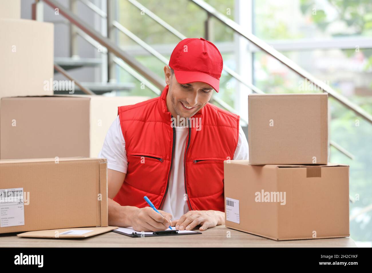 Young courier working with papers among parcels at table in delivery
