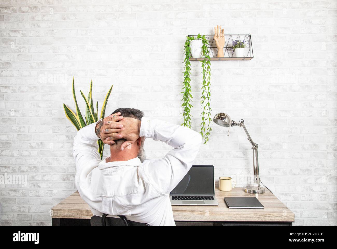 Relax in the Home Office. Rear view of freelancer resting at workplace, leaning on chair with hands behind head, working remotely. Stock Photo