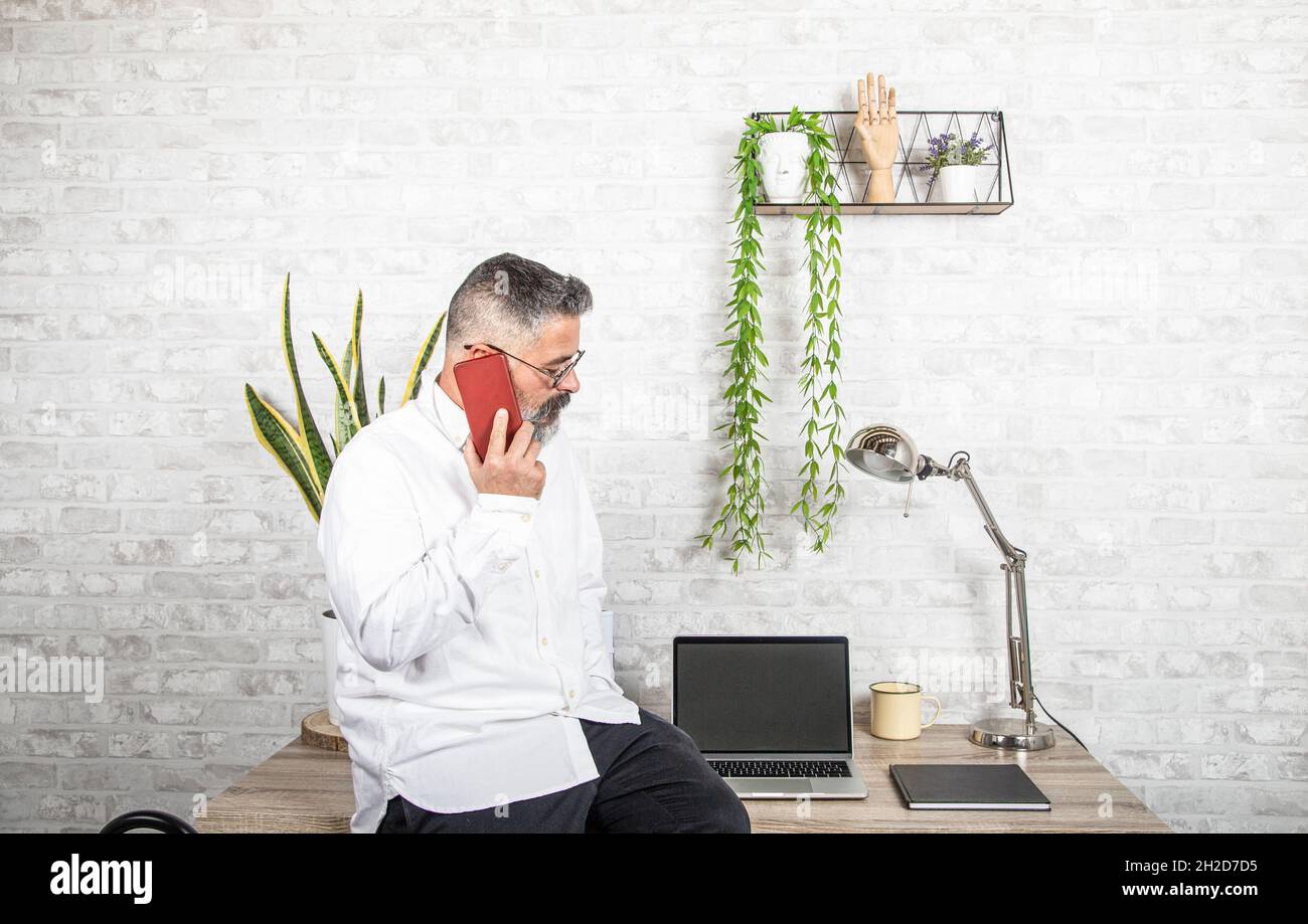 The guy works from home. He sits at the desk table and uses a laptop and talks on the phone. Stock Photo