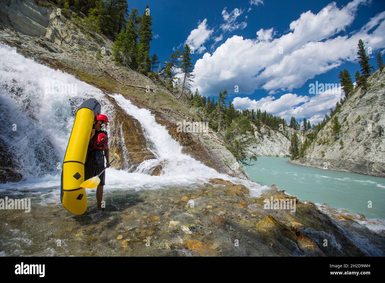 Paddler stands holding packraft next to waterfall, river. Stock Photo