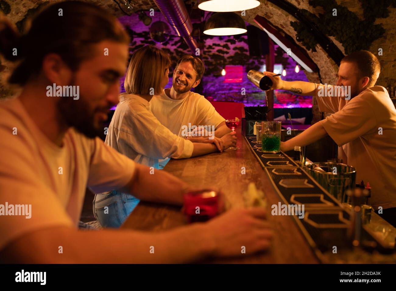 Adult couple chatting while bartender preparing cocktail near bearded man in pub Stock Photo