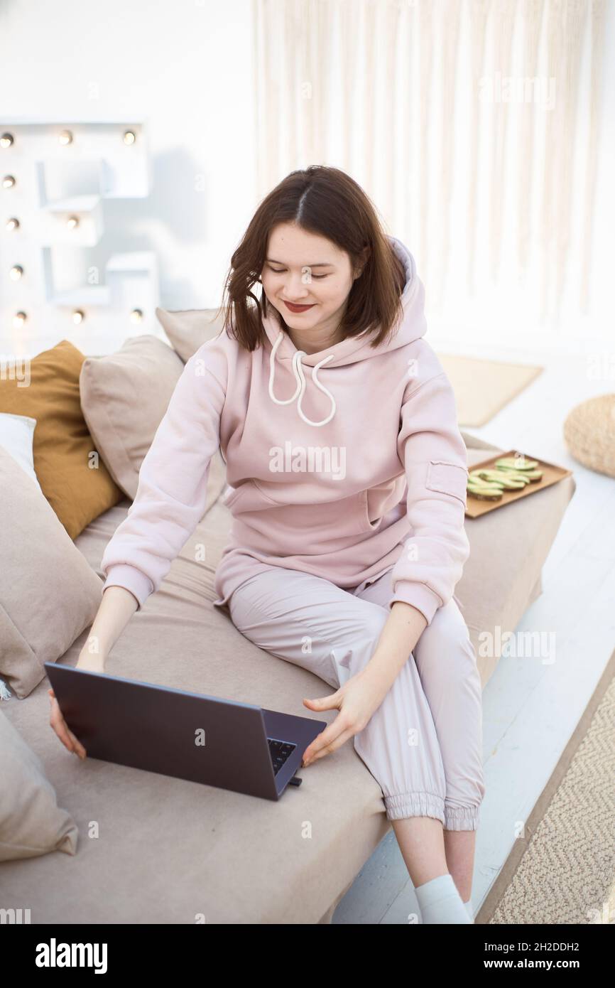 Young smiling woman is sitting on the couch and looking at the laptop Stock Photo