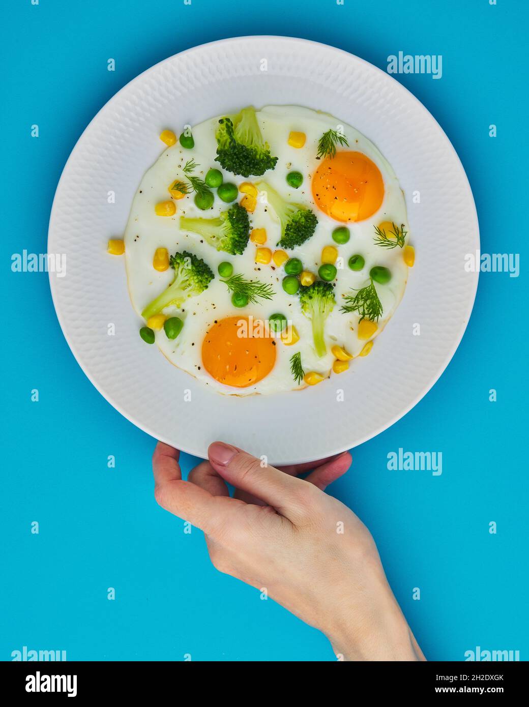 Female hand touching plate with fried eggs, broccoli, corn on blue