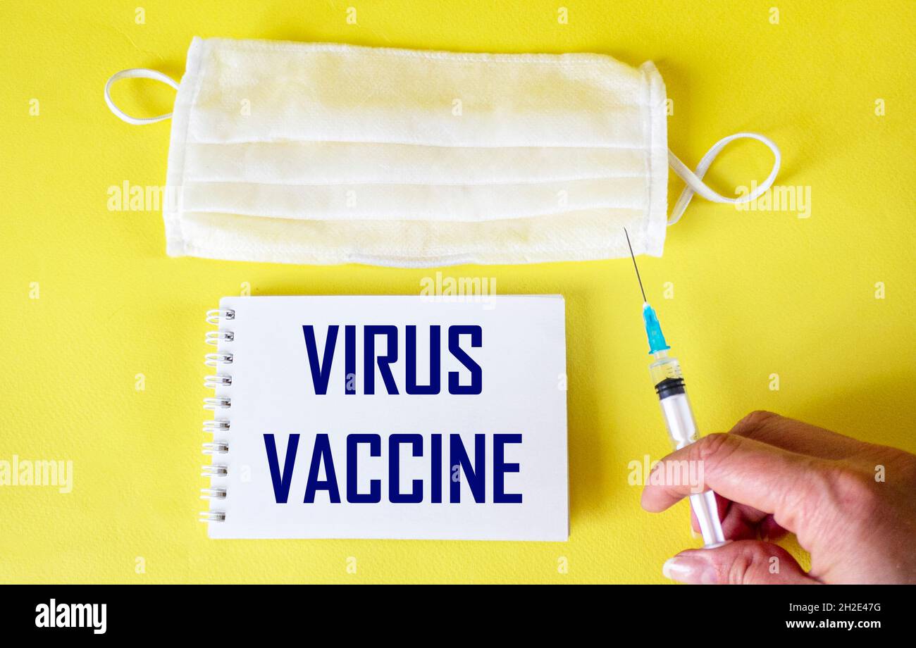 On a yellow background, a medina mask and a notebook with the text VIRUS VACCINE . Female hand with vaccine Stock Photo