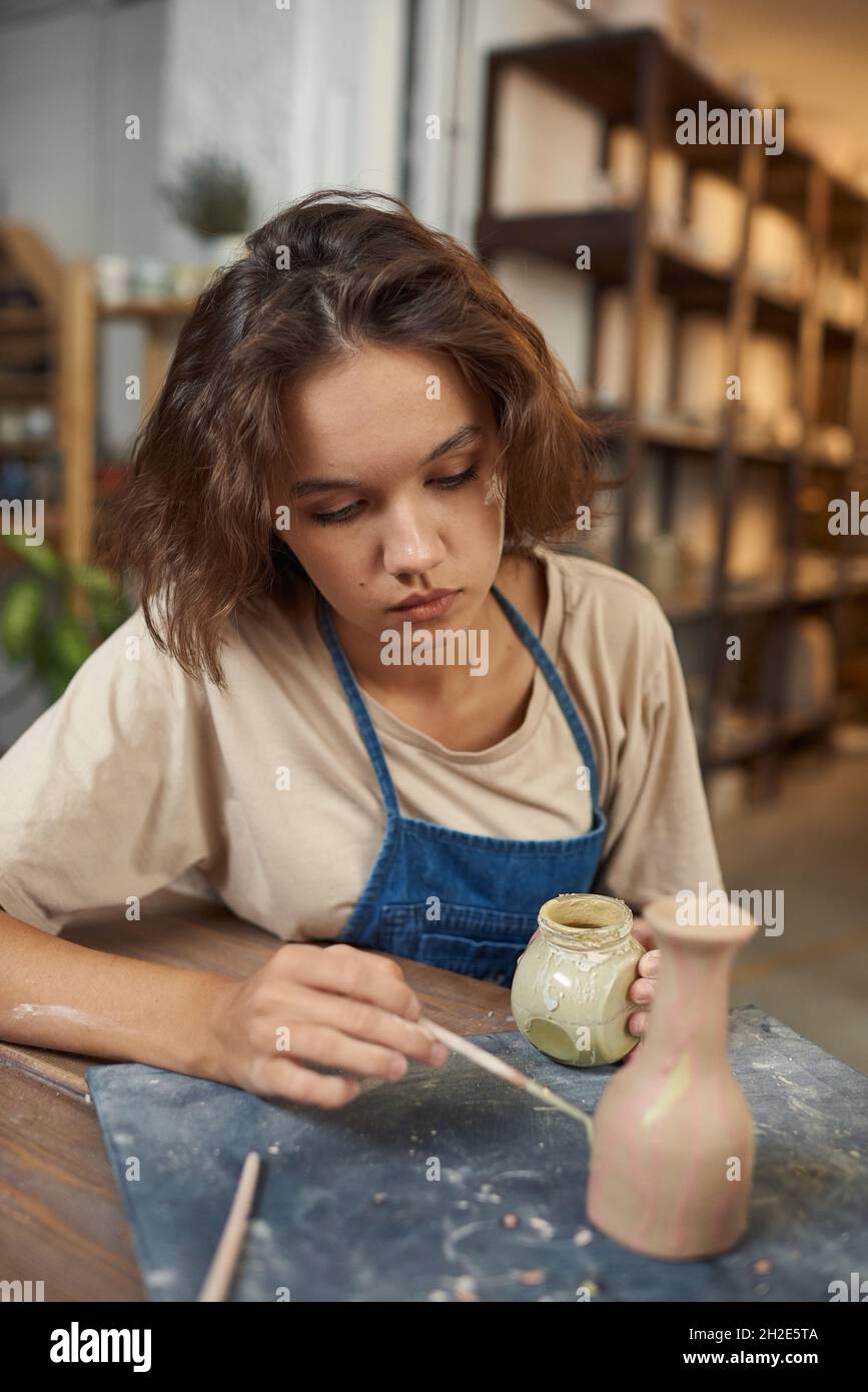 Concentrated young Caucasian potter sitting at table and painting vase while working on its design in workshop Stock Photo