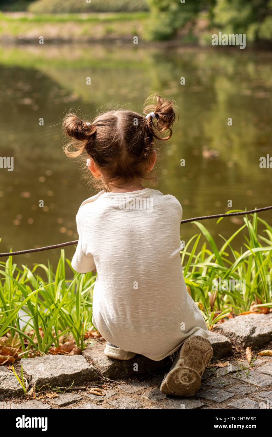 Little cute redhead girl with ponytails sits on the shore of a pond ...
