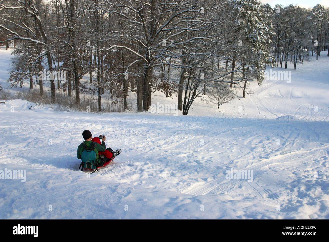 A father and child pile onto a plastic sled and take a selfie as they slide down a snowy hill in winter Stock Photo