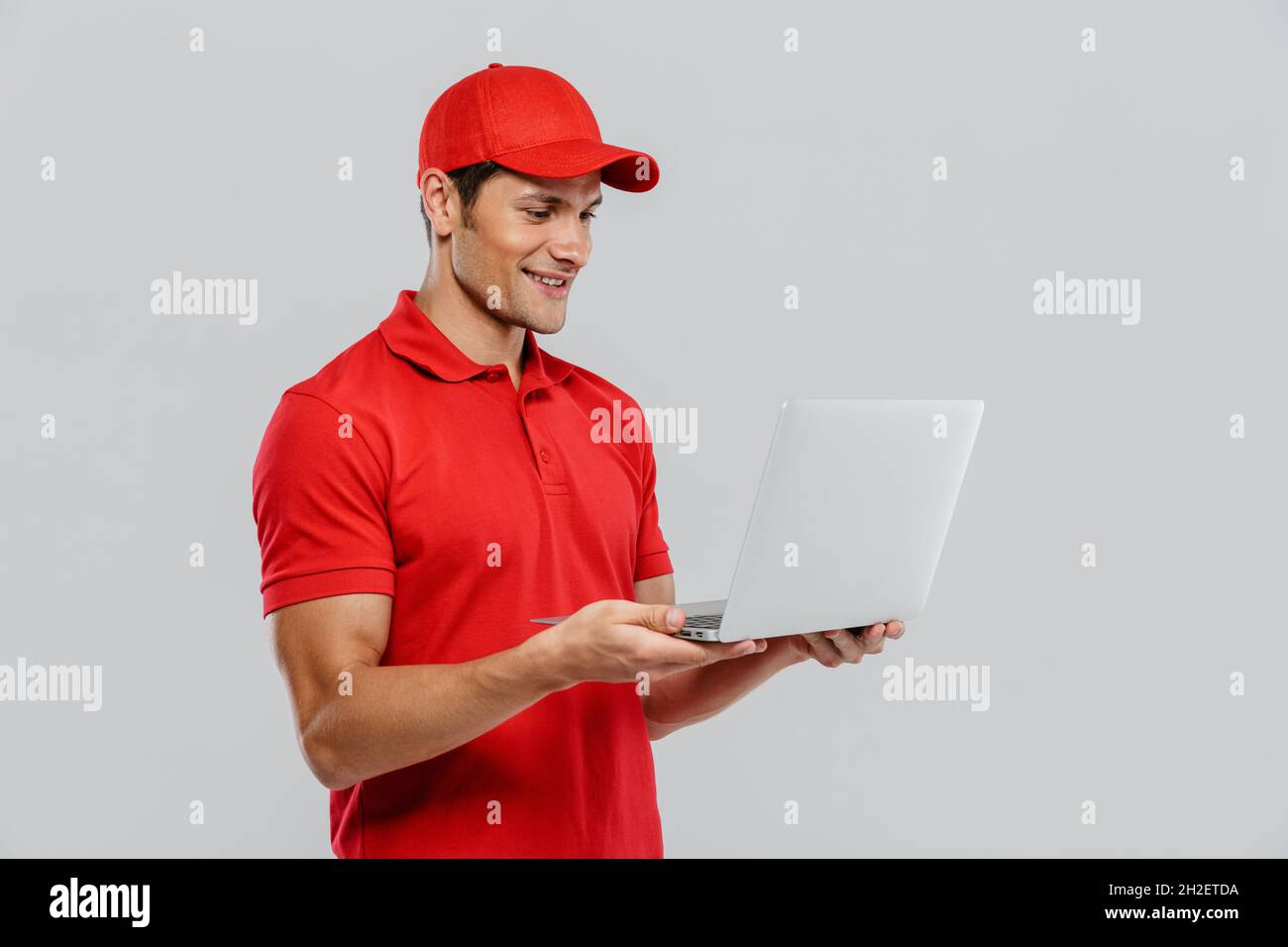 Young delivery man in uniform smiling while posing with laptop isolated ...