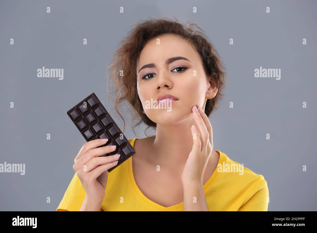 Beautiful young woman with acne problem holding chocolate on grey