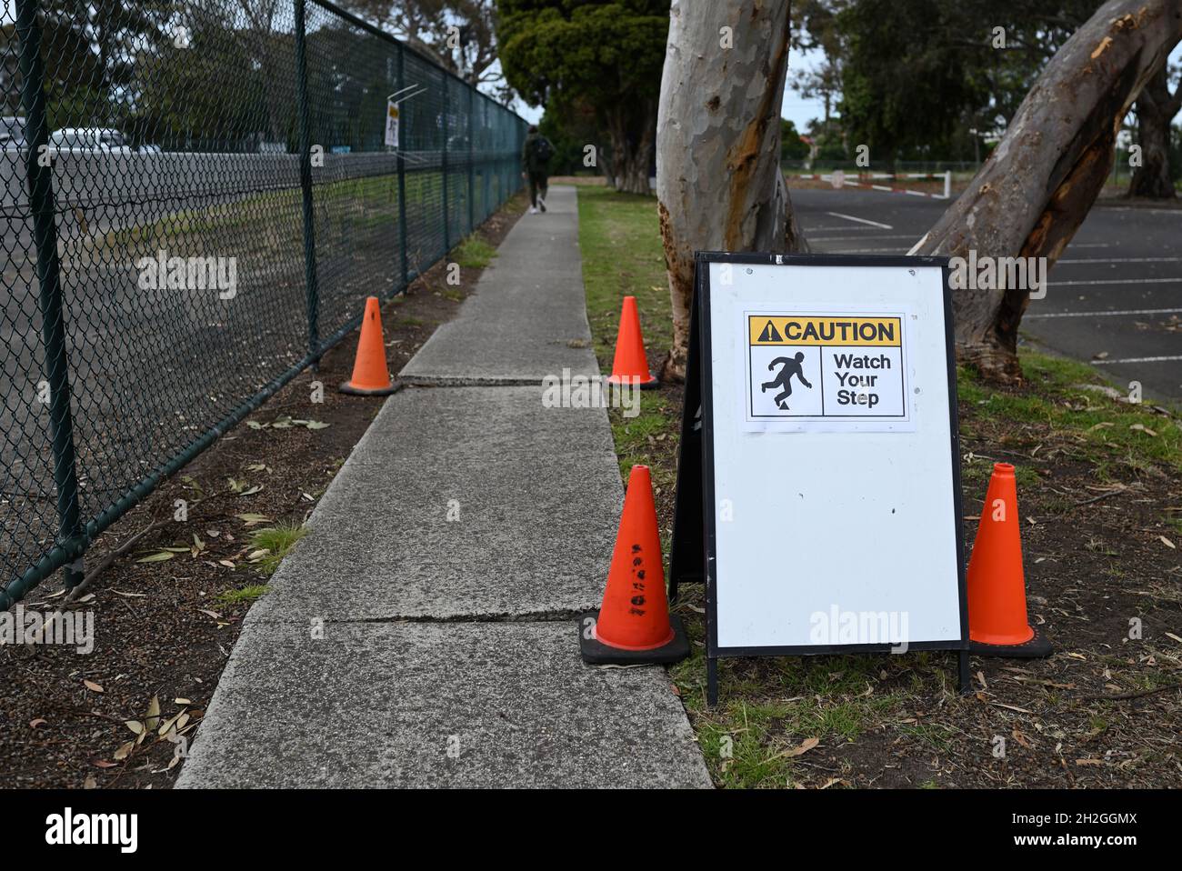 Caution watch your step sign, warning about uneven pavement on a footpath, with orange traffic cones. A person can be seen walking in the background Stock Photo