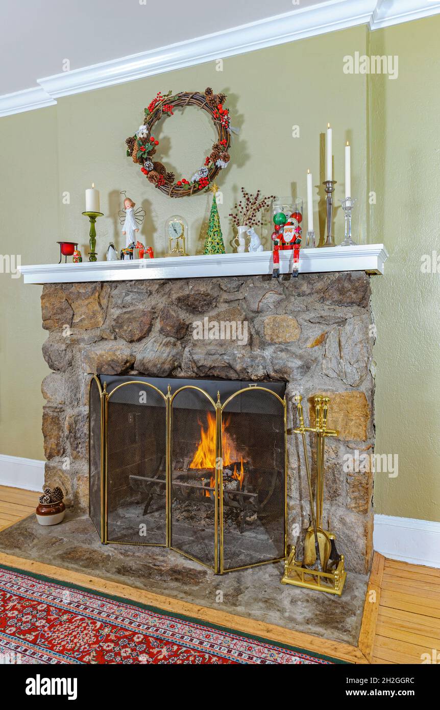 Cosy fieldstone fireplace with fire burning and mantelpiece decorated for Christmas with candles, Christmas decorations and a holiday wreath. Stock Photo