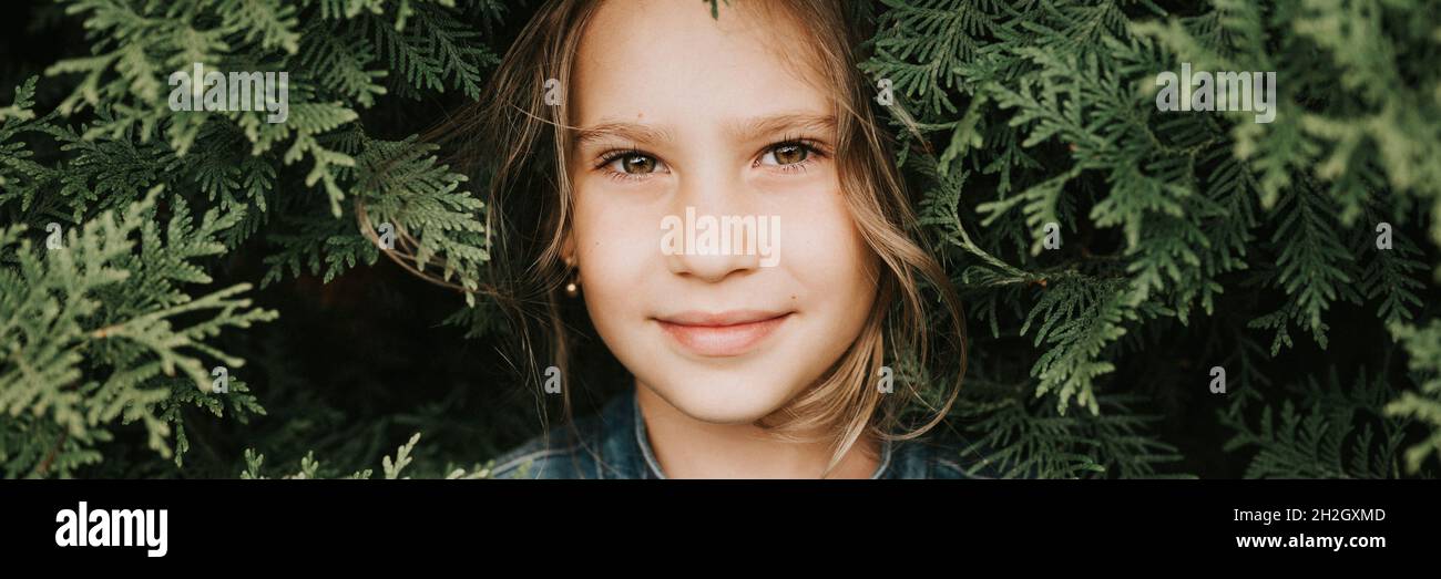portrait of the face of a cute happy caucasian candid healthy eight year old kid girl surrounded by branches and leaves of green plant thuja or cypres Stock Photo