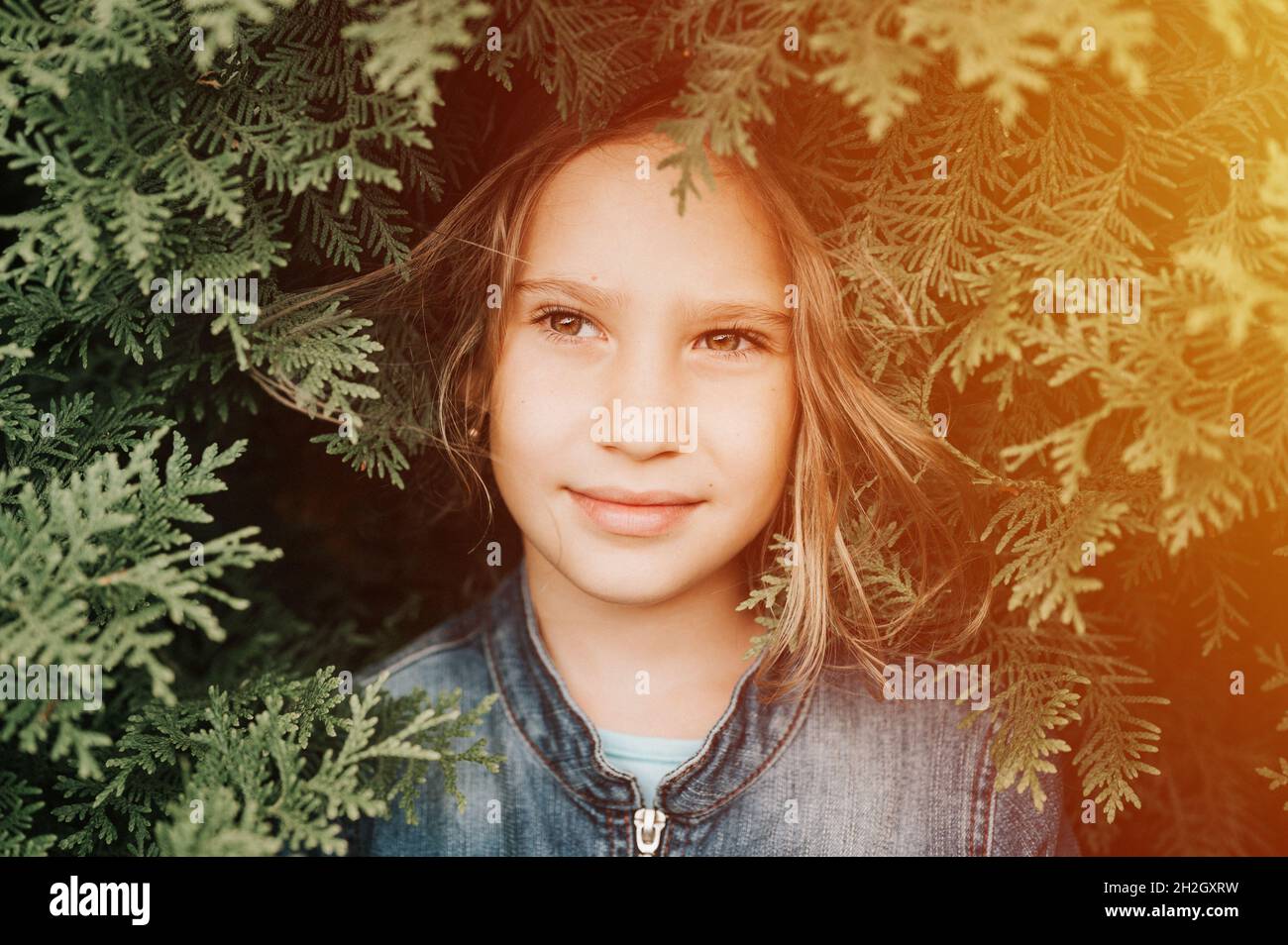 portrait of the face of a cute happy caucasian candid healthy eight year old kid girl surrounded by branches and leaves of green plant thuja or cypres Stock Photo