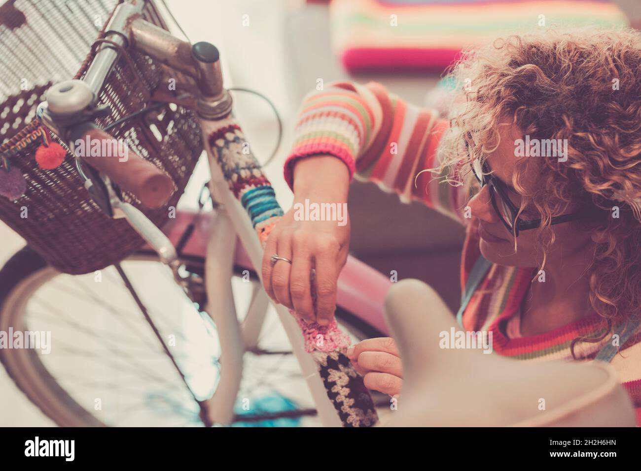 Woman decorating bicycle rod with woolen fabric. Woman with curly hair and eyeglasses decorating or modifying her bicycle with knitted woolen fabric o Stock Photo