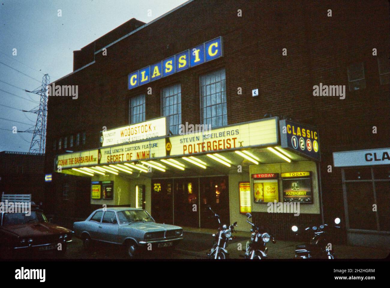 Classic Cinema, Hagley Road West, Quinton, Dudley, 1982. The Classic Cinema viewed from the south-east at dusk, showing the illuminated sign and billboard, with parked cars and motorcycles in front. The Danilo Cinema opened in 1939 and was the largest in Mortimer Dent's chain of Danilo Cinemas. It was later taken over by Essoldo, Classic, Cannon, ABC, Odeon, and finally by Reel Cinemas. In the early 21st century Reel Cinemas restored the building's facade to its original art deco style. Stock Photo