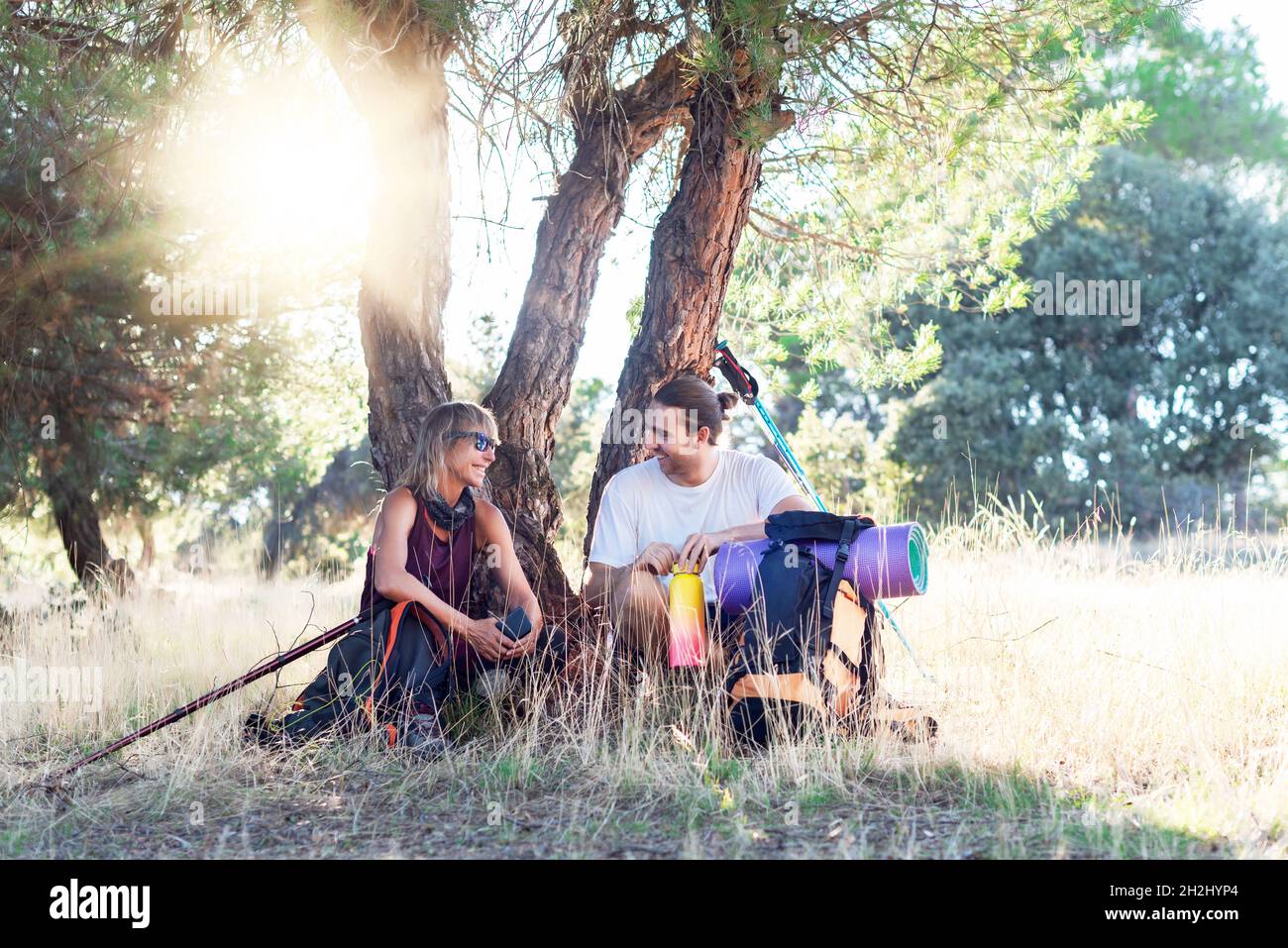 Two hikers, man and woman resting under the shade of a tree and chatting animatedly after a long walk. Stock Photo