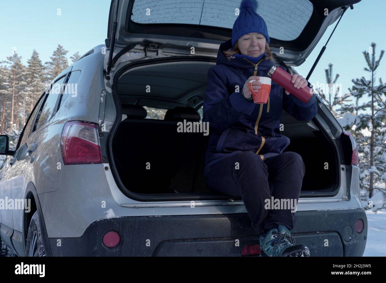Senior woman drinking tea while sitting in back of suv after wintry forest trail. Outdoor activities to boost mood during coronavirus pandemic at wint Stock Photo