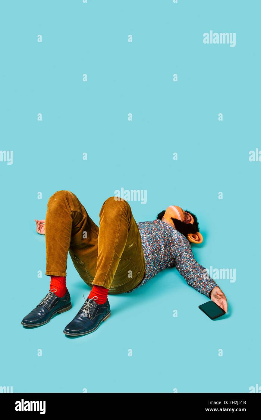 a young man, wearing a monkey mask, with his smartphone in his hand is lying down on a blue background with some blank space on top Stock Photo